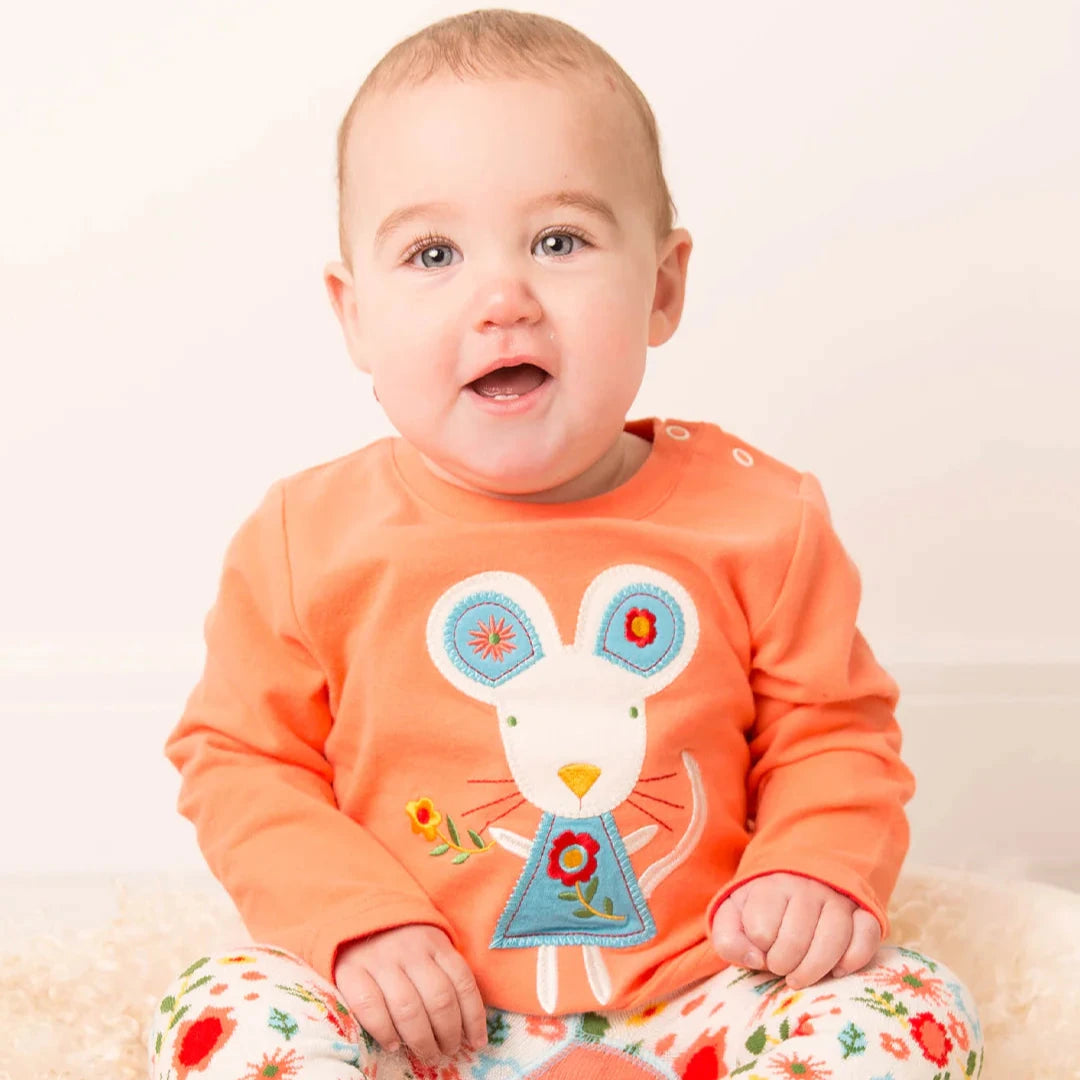 Baby wearing an orange shirt with a mouse design and floral pants sitting on a white surface.