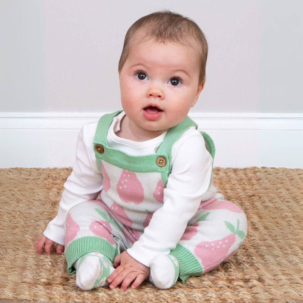 Baby wearing a white long-sleeve shirt with pink strawberry pattern and green overalls sitting on a textured surface.