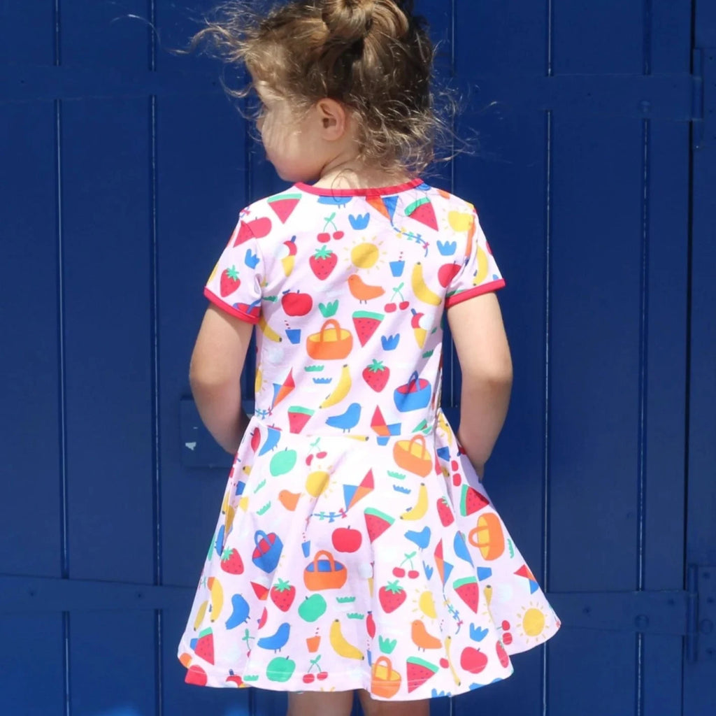 Child wearing a colorful dress with fruit pattern standing against a blue wall.