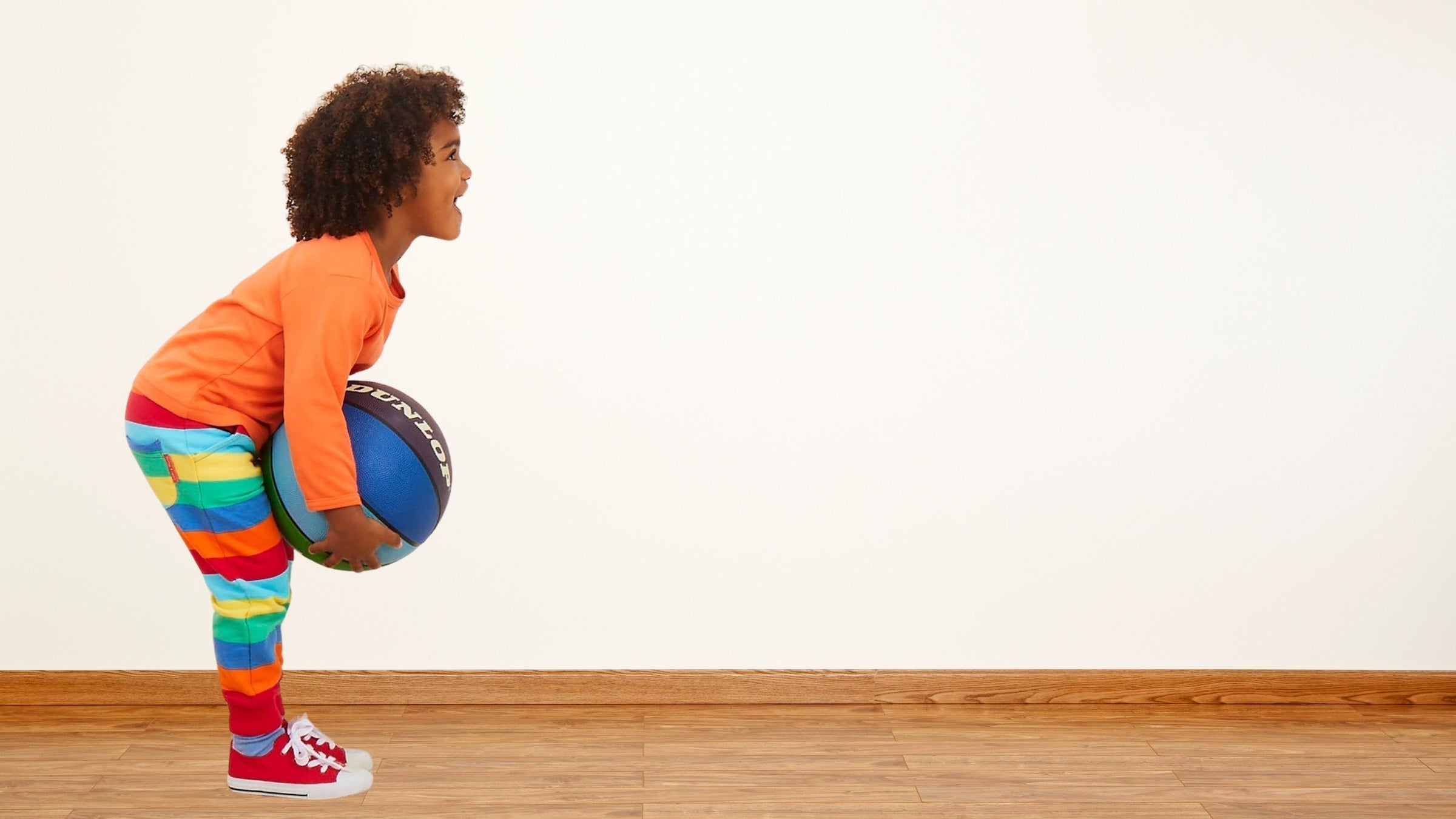 Vintage newborn clothes on a Child holding a basketball in a room with a white wall and wooden floor.
