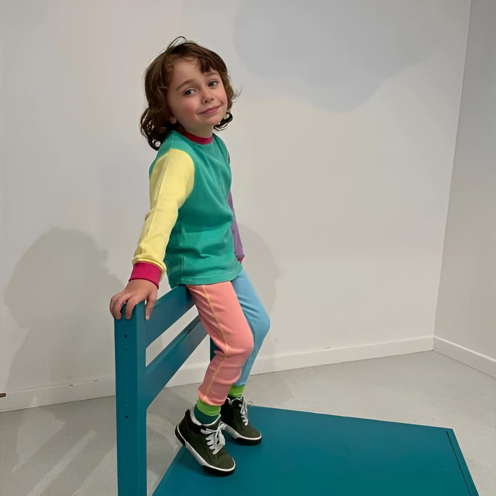 Child in colorful outfit standing on a blue chair against a white wall.