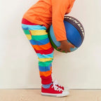 Child holding a basketball wearing an orange shirt and colorful striped pants against a white wall.