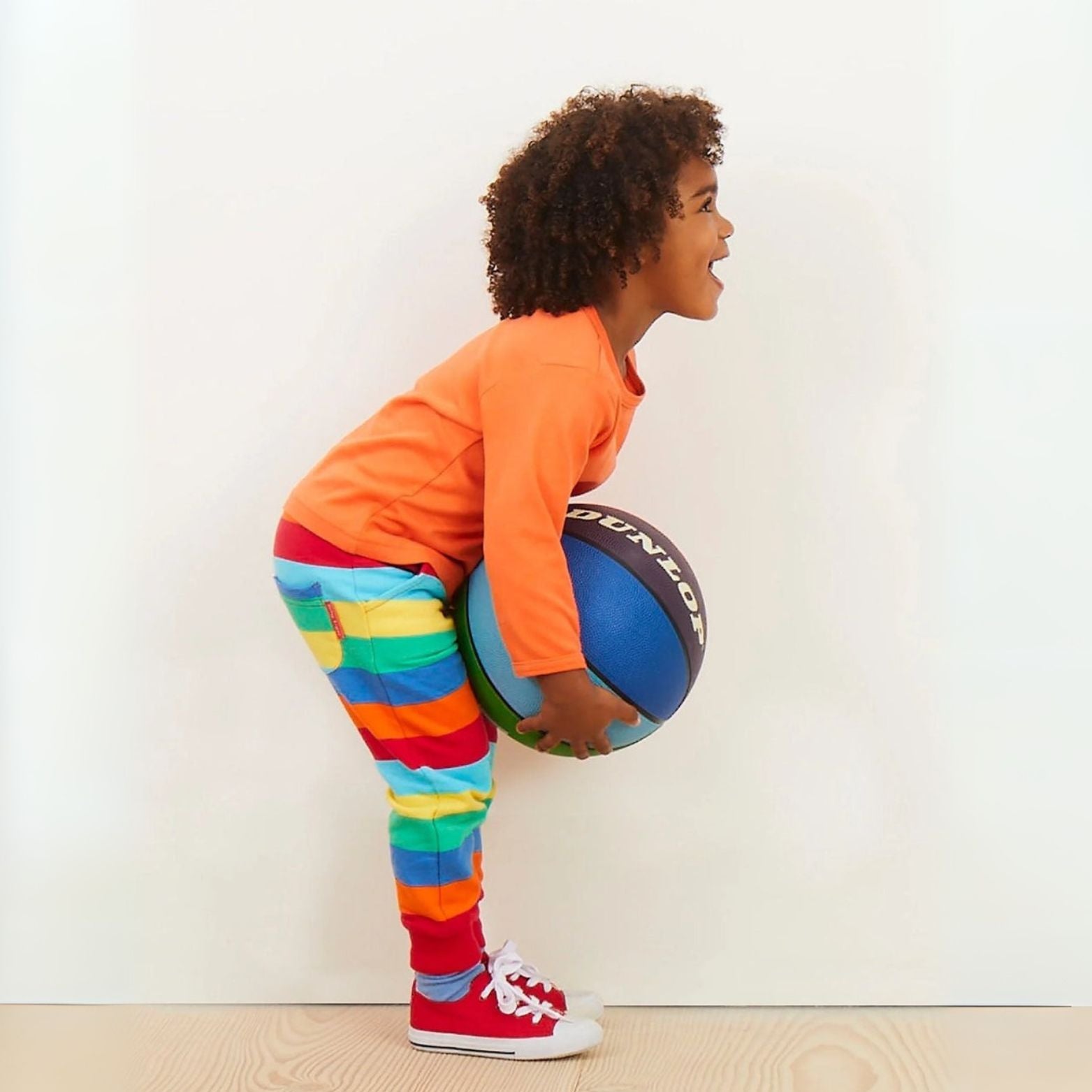 Child holding a basketball wearing an orange shirt and colorful striped pants against a white wall.