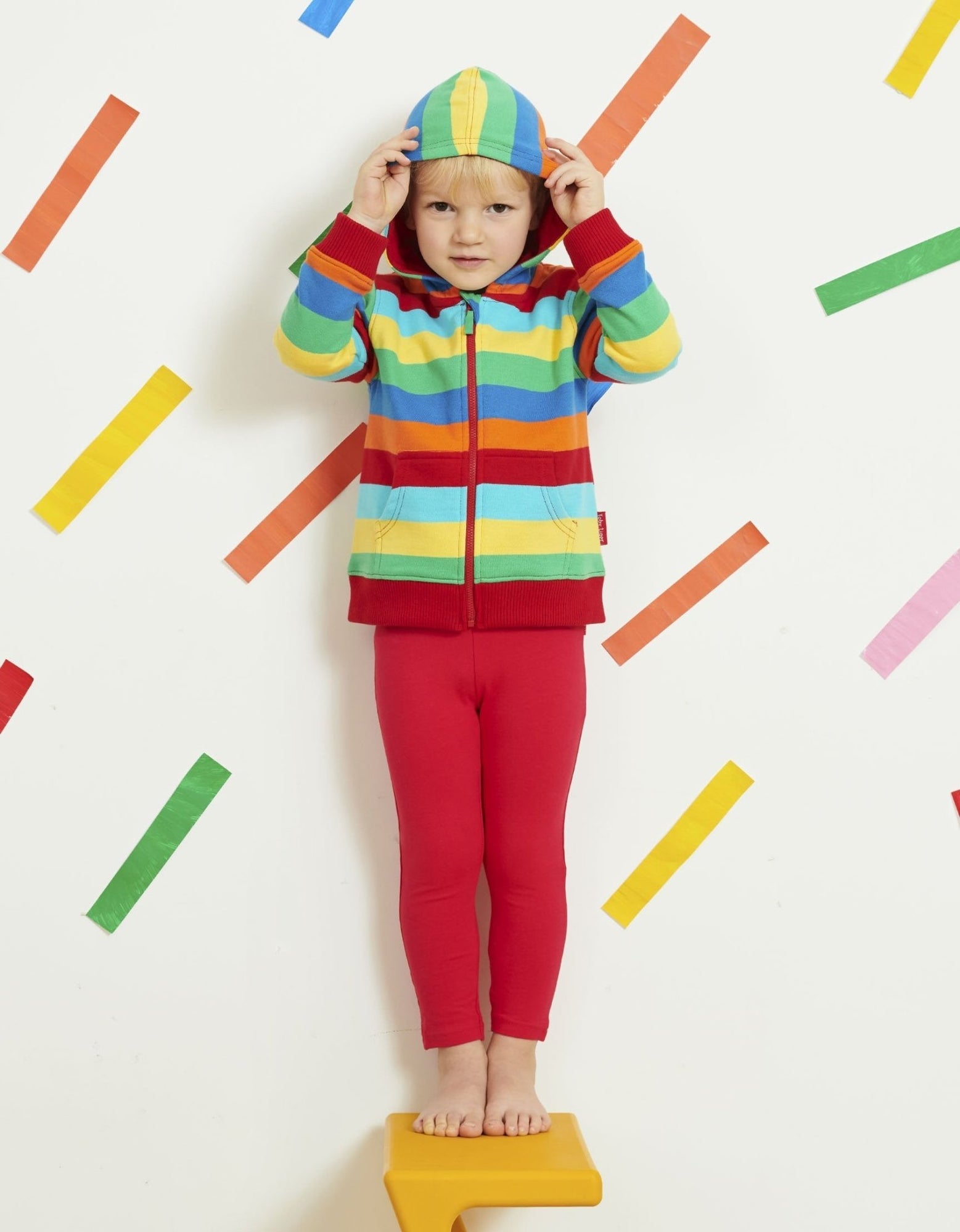 Child wearing a colorful striped hoodie and red pants standing on a yellow step against a white background with colorful bars.
