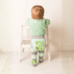 Baby wearing green and gray dinosaur-themed pants with a white chair in the background