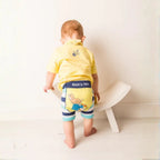 Child wearing a yellow shirt and striped shorts with a character design, standing on a white stool.