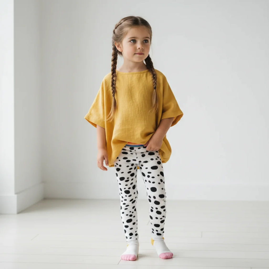 Young girl wearing a yellow top and polka dot leggings standing against a white background