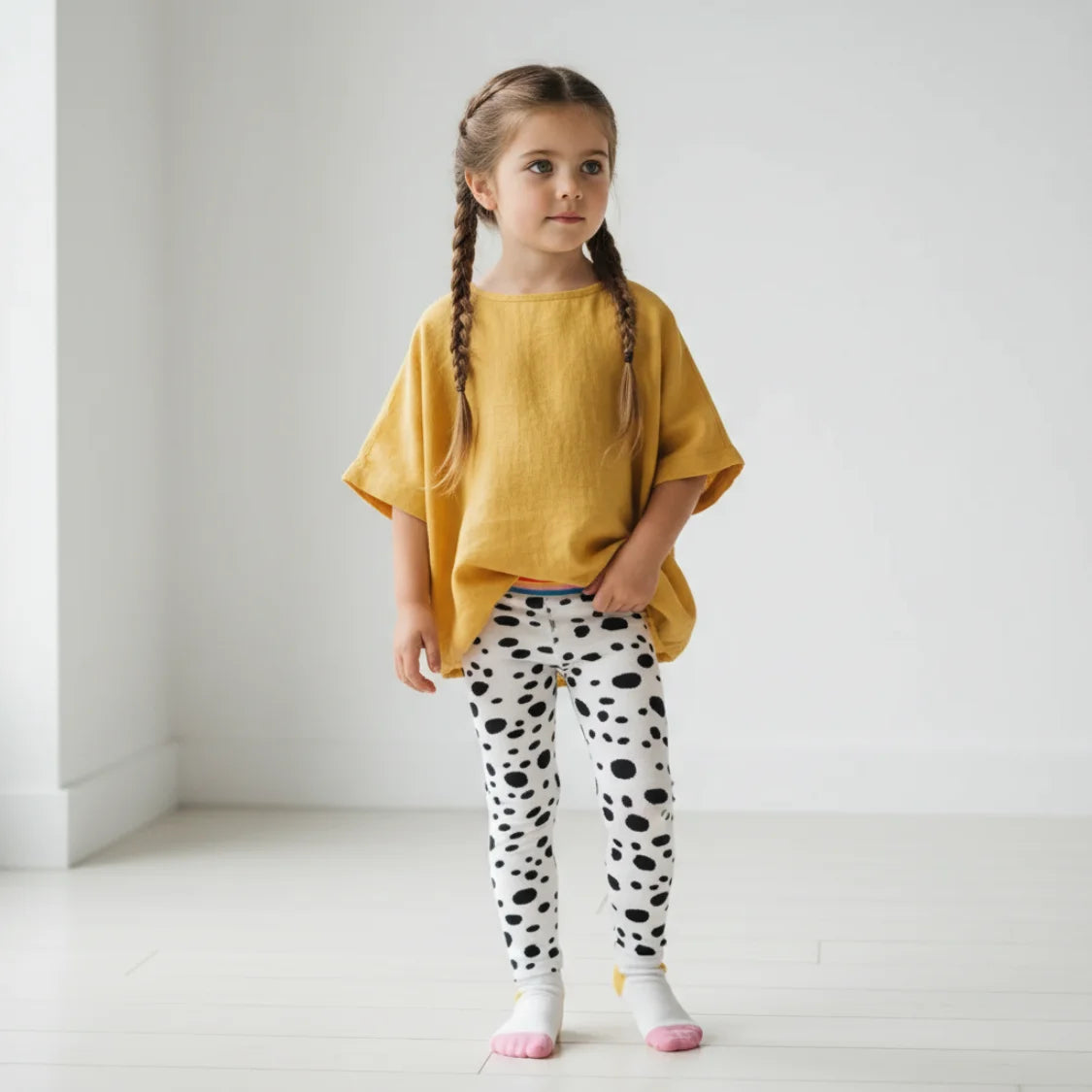 Young girl wearing a yellow top and polka dot leggings standing against a white background