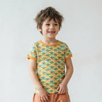 A young child standing in a bright room, wearing a short-sleeve t-shirt with a green and orange geometric pattern and loose orange pants.