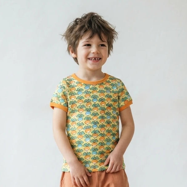 A young child standing in a bright room, wearing a short-sleeve t-shirt with a green and orange geometric pattern and loose orange pants.