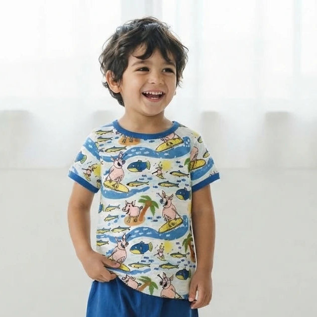 A young boy smiling and posing in a matching two-piece children's outfit. The set features a short-sleeve t-shirt with a colorful cartoon animal and wave pattern on a cream background, paired with solid blue jogger pants and white sneakers, set against a bright, minimalist white background.