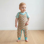 A baby standing on a light wood floor against a white wall, wearing a short-sleeve one-piece jumpsuit with a colorful fish print and light blue trim.