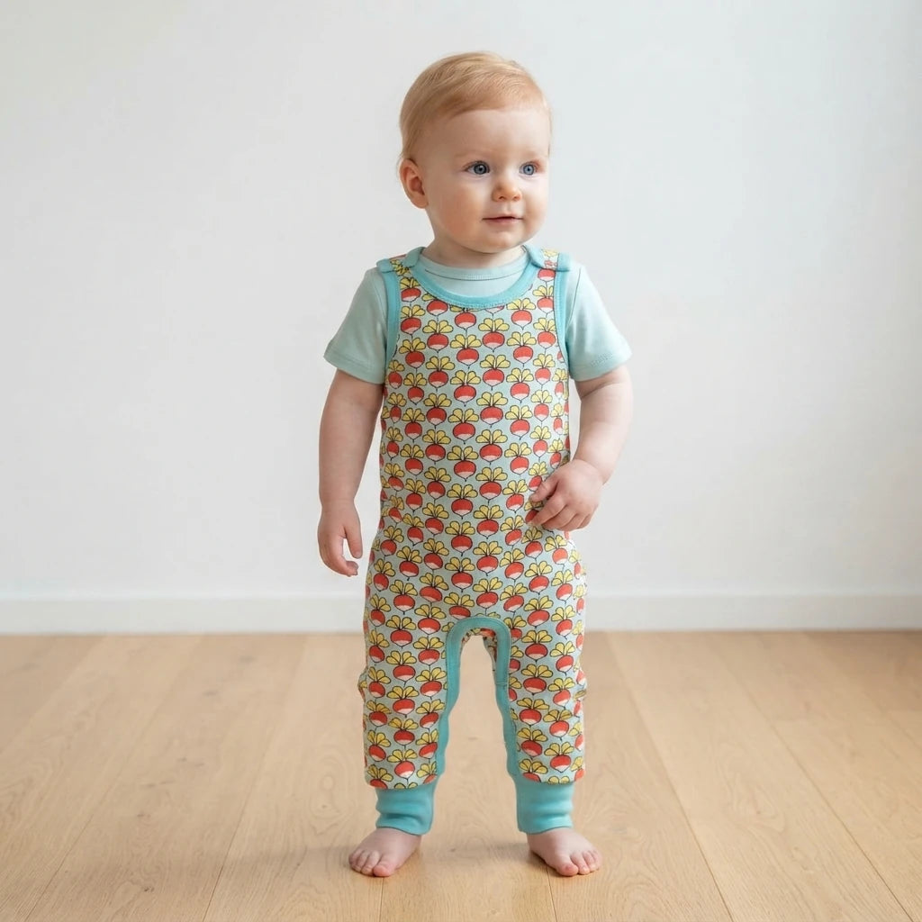 A baby standing on a light wood floor against a white wall, wearing a short-sleeve one-piece jumpsuit with a colorful fish print and light blue trim.