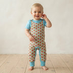 A smiling toddler standing barefoot on a light wood floor against a neutral cream wall, wearing a light blue short-sleeve top and matching light blue leg warmers over a one-piece romper with a colorful repeating pattern.