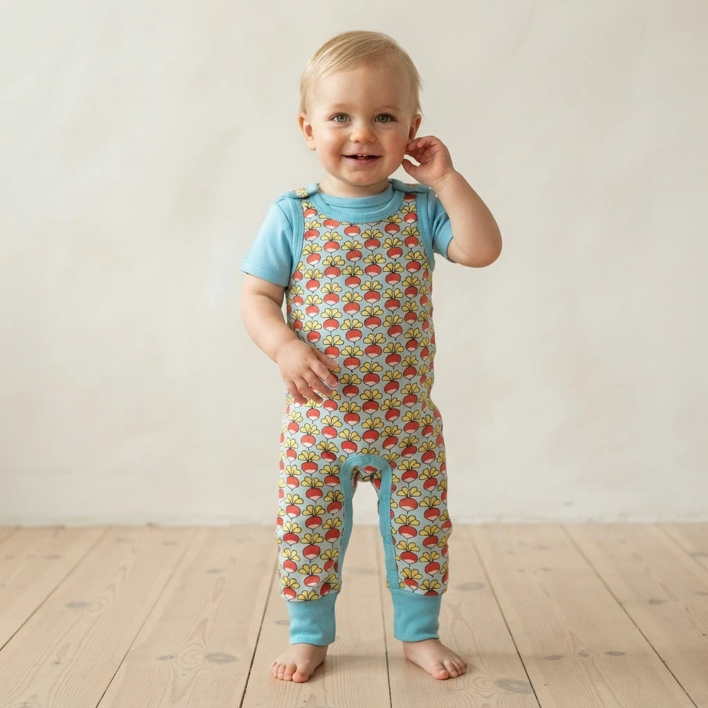 A smiling toddler standing barefoot on a light wood floor against a neutral cream wall, wearing a light blue short-sleeve top and matching light blue leg warmers over a one-piece romper with a colorful repeating pattern.
