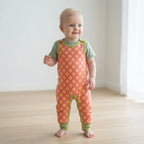 A baby standing barefoot on a light wood floor wearing a pink and yellow patterned footless overalls romper with sage green sleeves and leg cuffs.