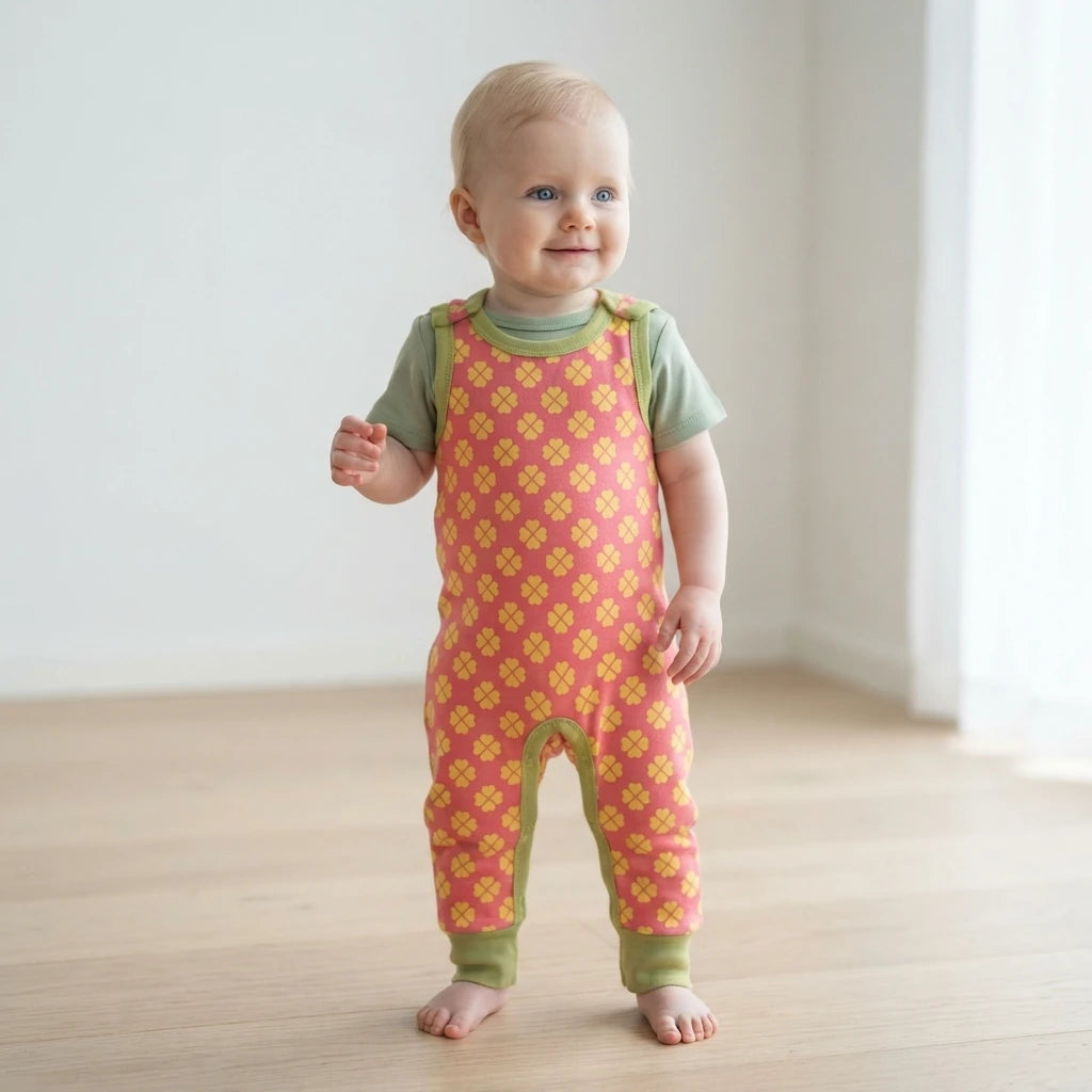 A baby standing barefoot on a light wood floor wearing a pink and yellow patterned footless overalls romper with sage green sleeves and leg cuffs.