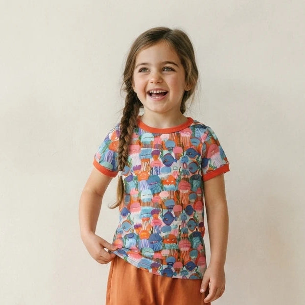A young girl with braided hair stands smiling in a studio setting, wearing a short-sleeve ringer t-shirt with a colorful abstract pattern and orange trim, paired with orange pants and white sneakers.