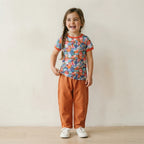 A young girl with braided hair stands smiling in a studio setting, wearing a short-sleeve ringer t-shirt with a colorful abstract pattern and orange trim, paired with orange pants and white sneakers.