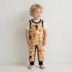 A toddler with curly blonde hair stands barefoot against a plain white wall, wearing a short-sleeve floral jumpsuit over a cream t-shirt. The jumpsuit features a vibrant tropical print with toucans, watermelons, and hibiscus flowers, accented by black trim.