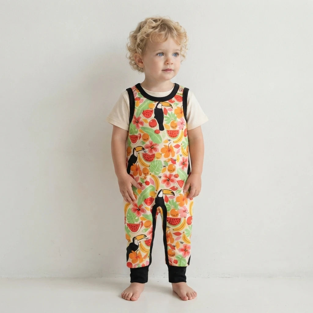 A toddler with curly blonde hair stands barefoot against a plain white wall, wearing a short-sleeve floral jumpsuit over a cream t-shirt. The jumpsuit features a vibrant tropical print with toucans, watermelons, and hibiscus flowers, accented by black trim.