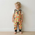 A toddler with curly blonde hair stands barefoot against a plain off-white wall, wearing a short-sleeve jumpsuit with a colorful tropical print of toucans, flowers, and fruit, layered over a cream t-shirt.