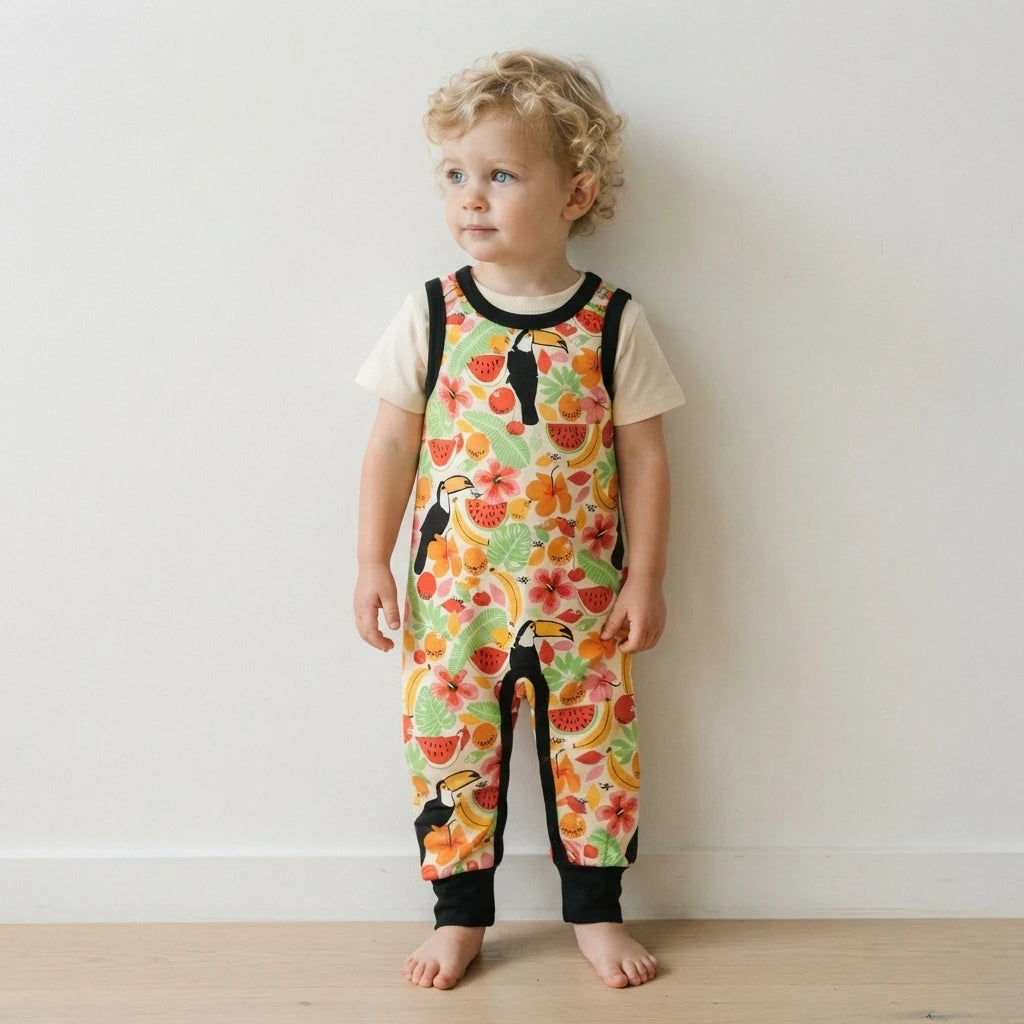 A toddler with curly blonde hair stands barefoot against a plain off-white wall, wearing a short-sleeve jumpsuit with a colorful tropical print of toucans, flowers, and fruit, layered over a cream t-shirt.