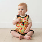 A baby sitting on a light wood floor against a white wall, wearing a short-sleeve romper with a colorful tropical print of toucans, fruit, and flowers over a cream shirt.