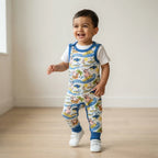 A smiling toddler walking indoors wearing a white short-sleeve shirt under a blue sleeveless overall-style romper with a colorful cartoon animal print, paired with blue ribbed cuffs and white sneakers.