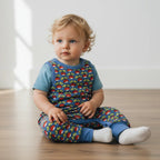 baby wearing a colorful dungarees with a blue shirt and patterned overalls sitting on a wooden floor.