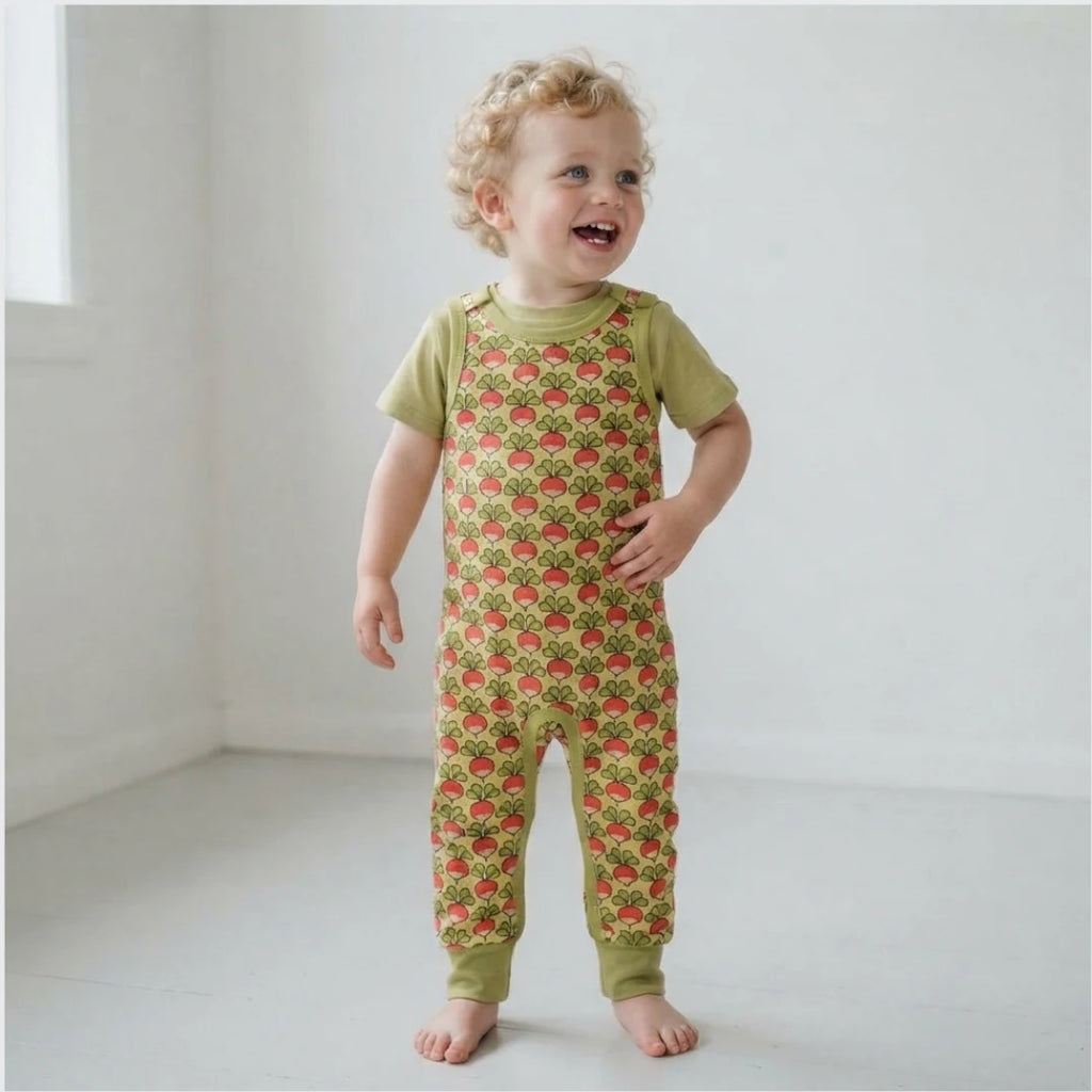 A toddler with curly blonde hair stands barefoot in a bright white room, wearing a short-sleeve green jumpsuit covered in a red strawberry print.