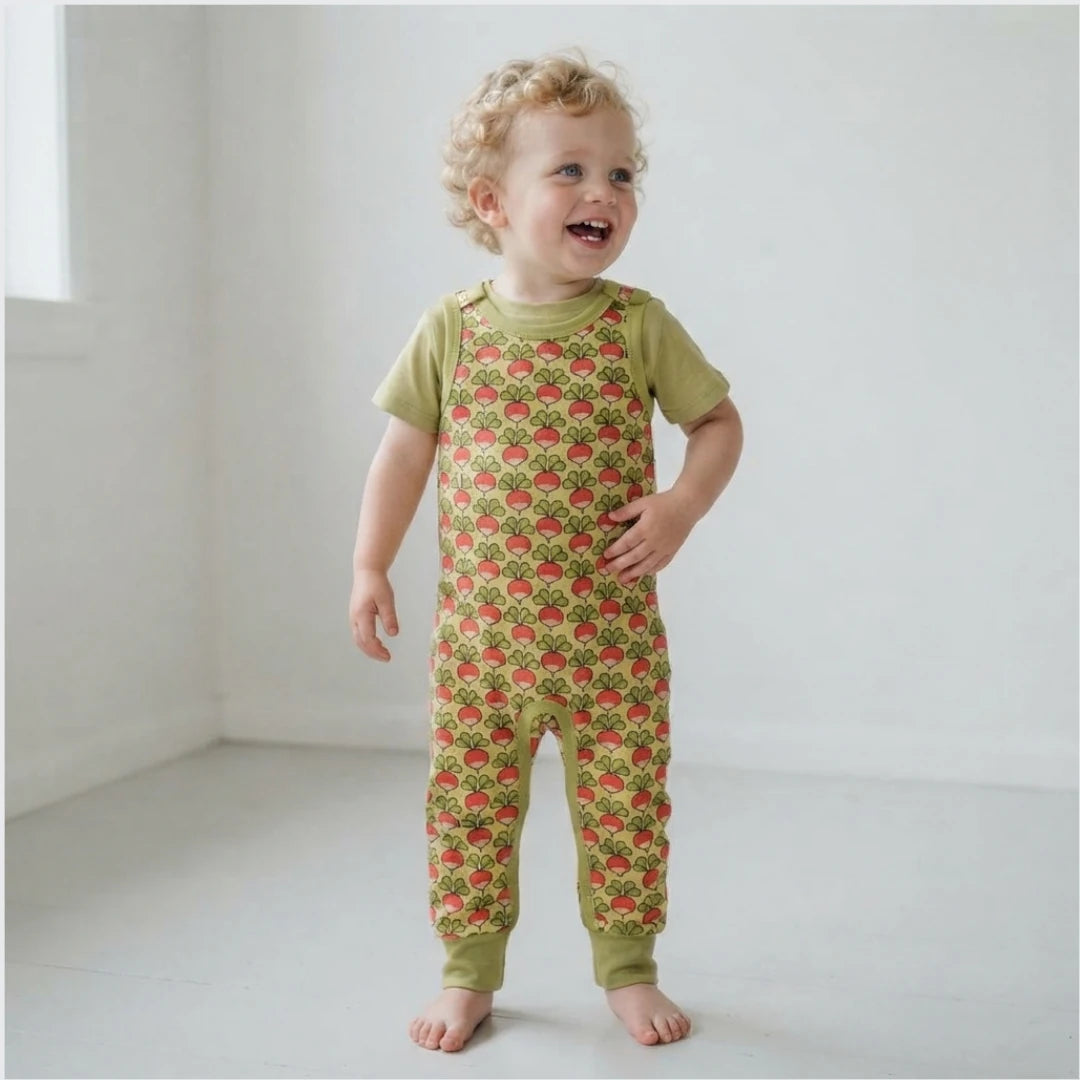 A toddler with curly blonde hair stands barefoot in a bright white room, wearing a short-sleeve green jumpsuit covered in a red strawberry print.