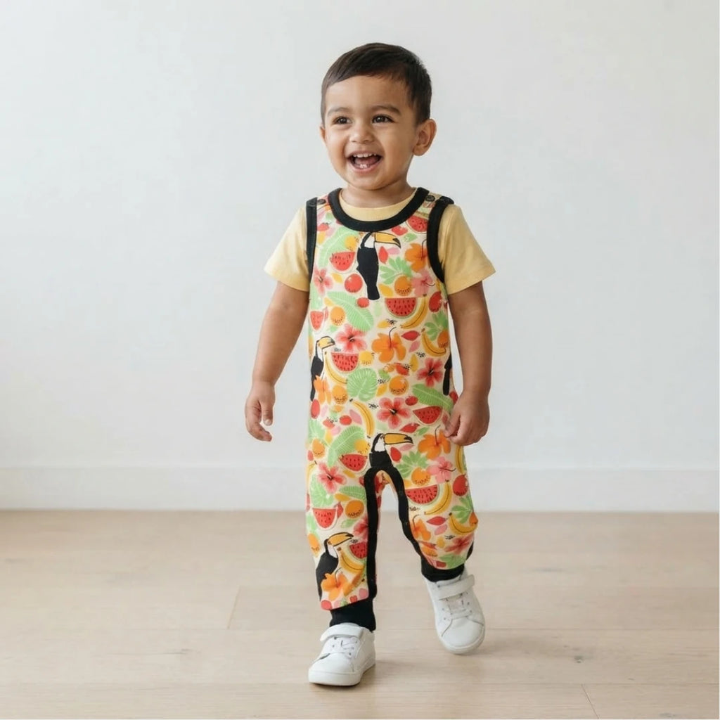 A smiling toddler walking indoors wearing a short-sleeve overall jumpsuit with a colorful tropical fruit and toucan print over a pale yellow shirt, paired with white sneakers.