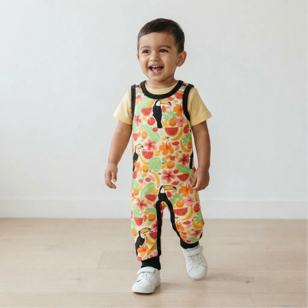 A smiling toddler walking indoors wearing a short-sleeve overall jumpsuit with a colorful tropical fruit and toucan print over a pale yellow shirt, paired with white sneakers.
