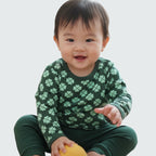 Child wearing a green outfit with clover pattern holding a yellow object on a plain background