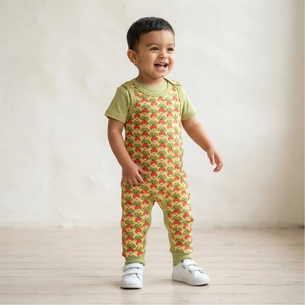A toddler standing in a studio wearing a light green short-sleeve jumpsuit with a red strawberry print over a light green shirt, paired with white shoes.
