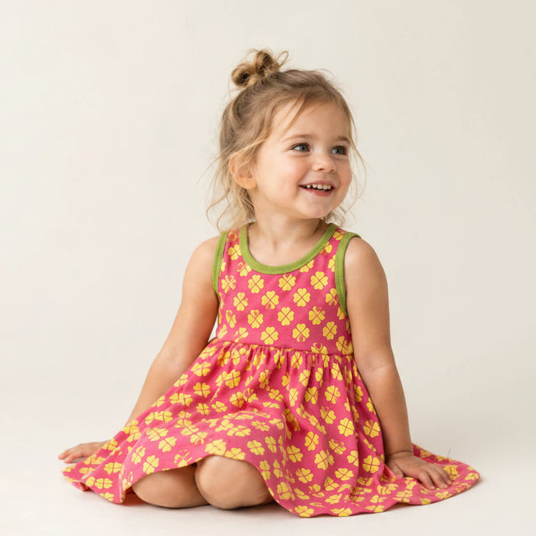 A young girl with a small bun sits on a white studio floor wearing a pink sleeveless dress with a yellow floral pattern and green trim.