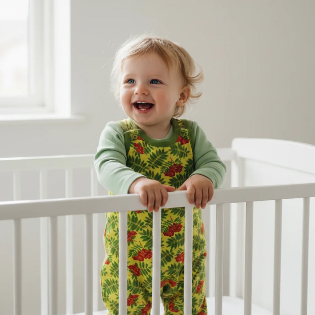 Child in a green shirt and yellow floral overalls standing in a crib.