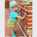 Baby wearing a colorful outfit standing against a brick wall with 'DUNS' branding.