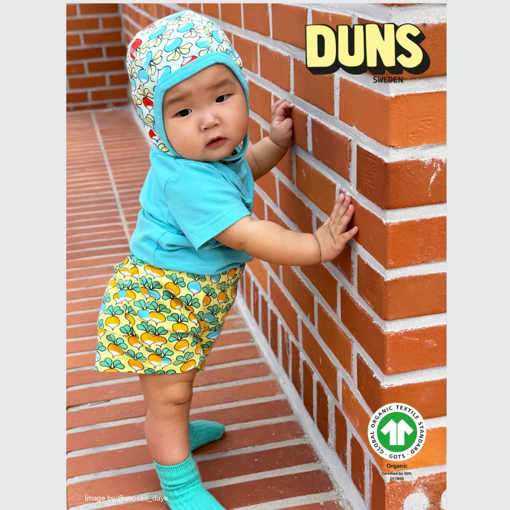 Baby wearing a colorful outfit standing against a brick wall with 'DUNS' branding.