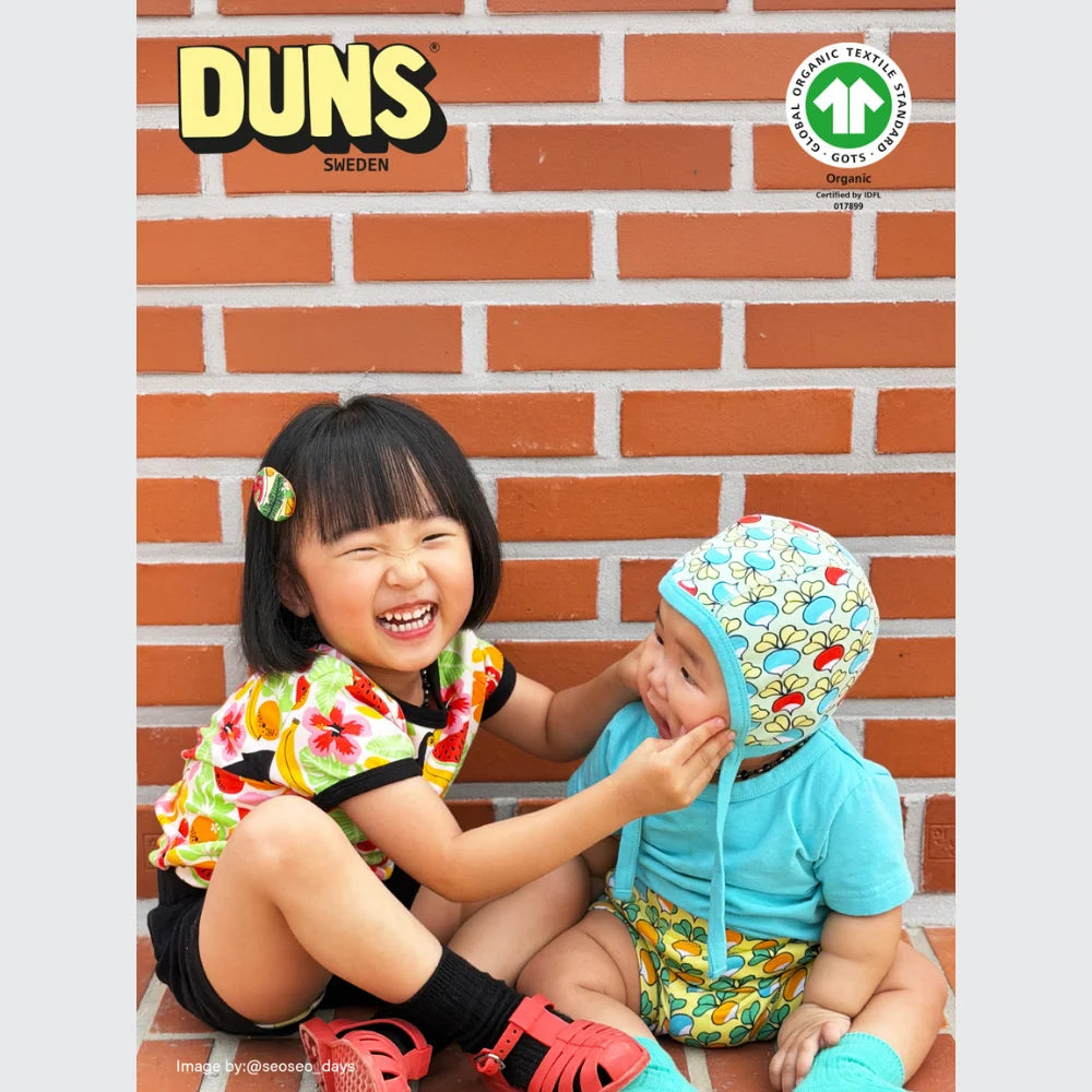 Two children wearing colorful outfits sitting against a brick wall with DUNS Sweden branding.