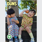 Two children in colorful outfits sitting on a bench with apples, featuring the brand 'DUNS'.