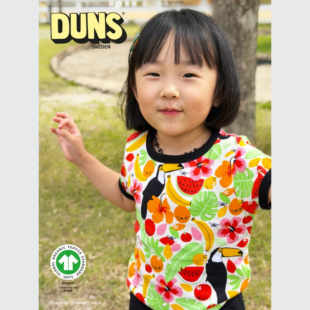 Child wearing a colorful shirt with fruit patterns outdoors, featuring the Duns Sweden logo.