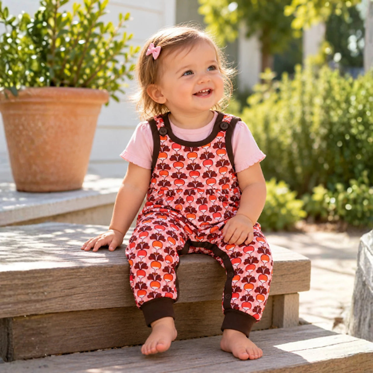 Toddler wearing the DUNS Sweden Radish Pink Dungarees in organic cotton, sitting on garden steps
