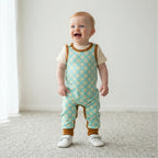 A baby standing on a light carpet in a bright room, wearing a short-sleeve overall romper with a mint green and cream floral pattern, brown trim, and white shoes.