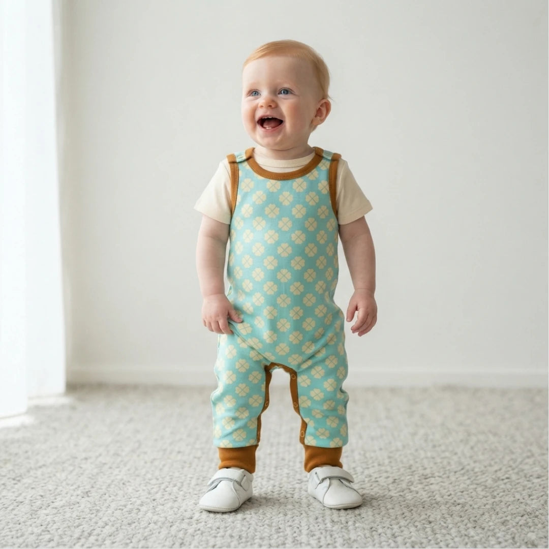 A baby standing on a light carpet in a bright room, wearing a short-sleeve overall romper with a mint green and cream floral pattern, brown trim, and white shoes.