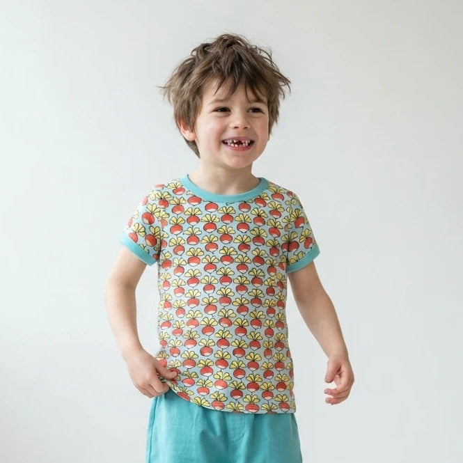 A young child standing in a bright room wearing a short-sleeve t-shirt with a colorful retro pattern and teal trim, paired with teal pants and white sneakers.