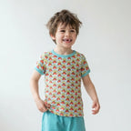 A young child standing in a bright room wearing a short-sleeve t-shirt with a colorful retro pattern and teal trim, paired with teal pants and white sneakers.