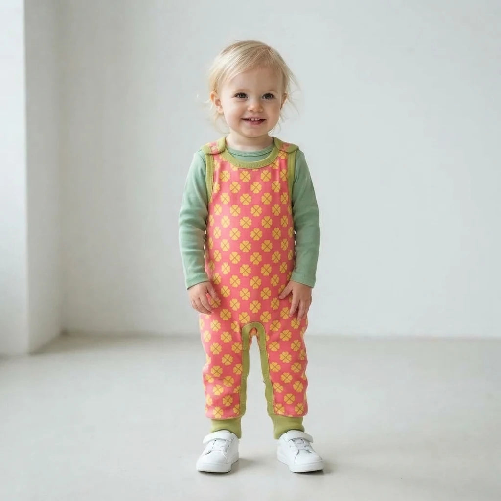 A toddler standing in a bright, minimalist room wearing a pink and yellow patterned overall jumpsuit over a sage green long-sleeve shirt and white sneakers.