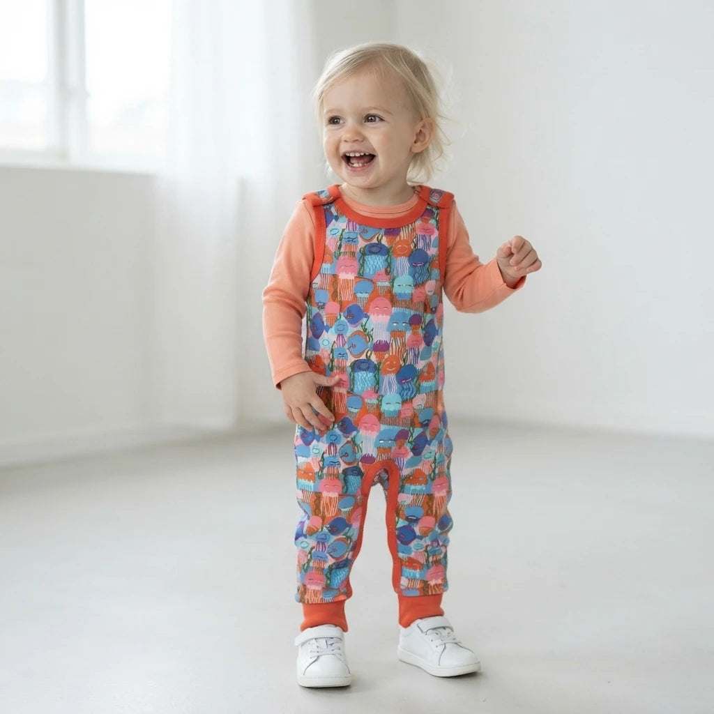 A toddler stands in a bright, minimalist white room wearing a long-sleeve orange shirt and colorful patterned baby dungarees with white sneakers.